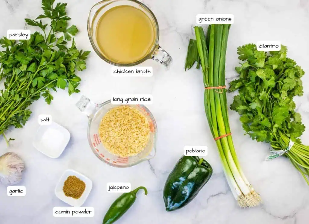 Assorted ingredients on a white surface, including parsley, cilantro, green onions, long grain rice, chicken broth, cumin powder, salt, garlic, jalapeno, and poblano peppers.