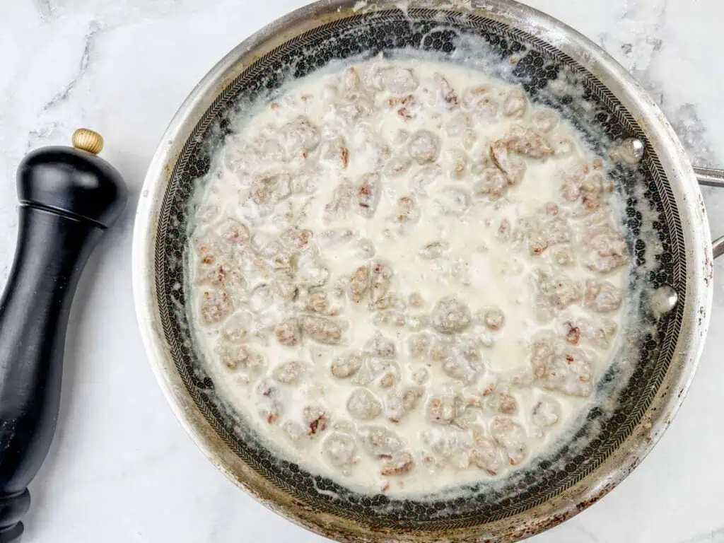 A skillet filled with cooked sausage in creamy white gravy, placed on a marble countertop with a black pepper grinder nearby.