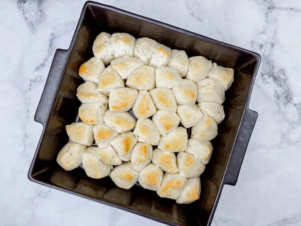 A square baking pan filled with evenly baked, golden-brown pull-apart bread rolls on a marble surface.