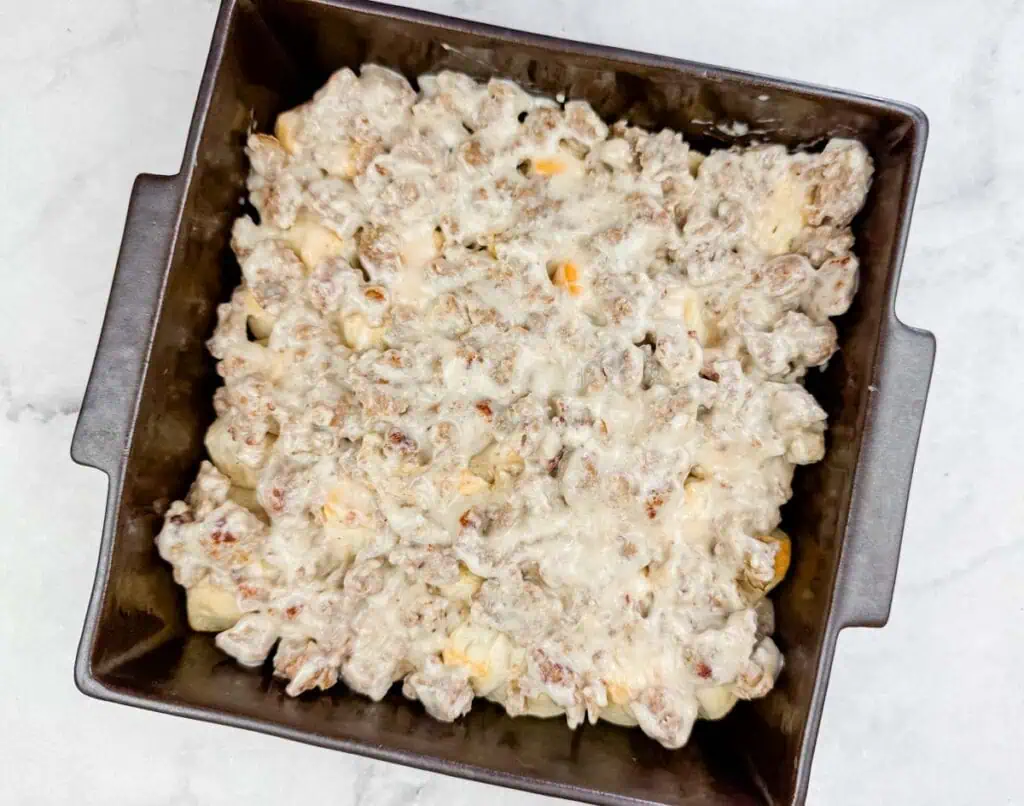 A square baking pan filled with a baked casserole topped with melted cheese and pieces of sausage on a light surface.