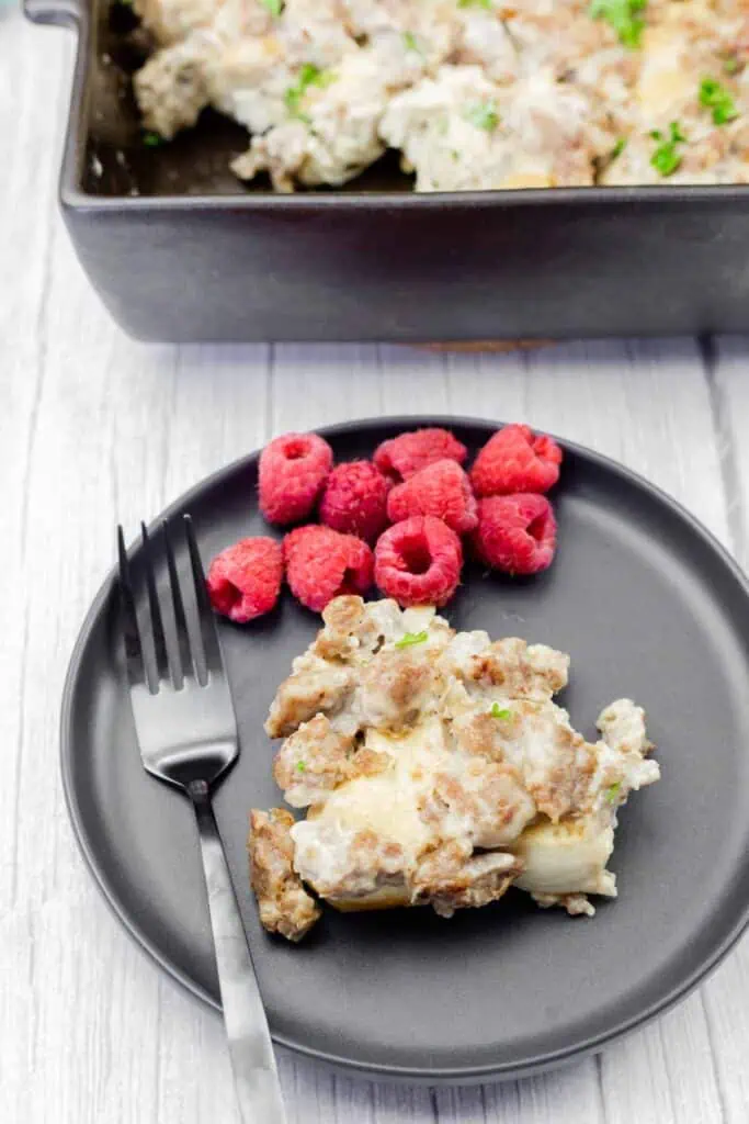 A black plate with a serving of sausage casserole and fresh raspberries, alongside a black fork. A casserole dish is partially visible in the background.