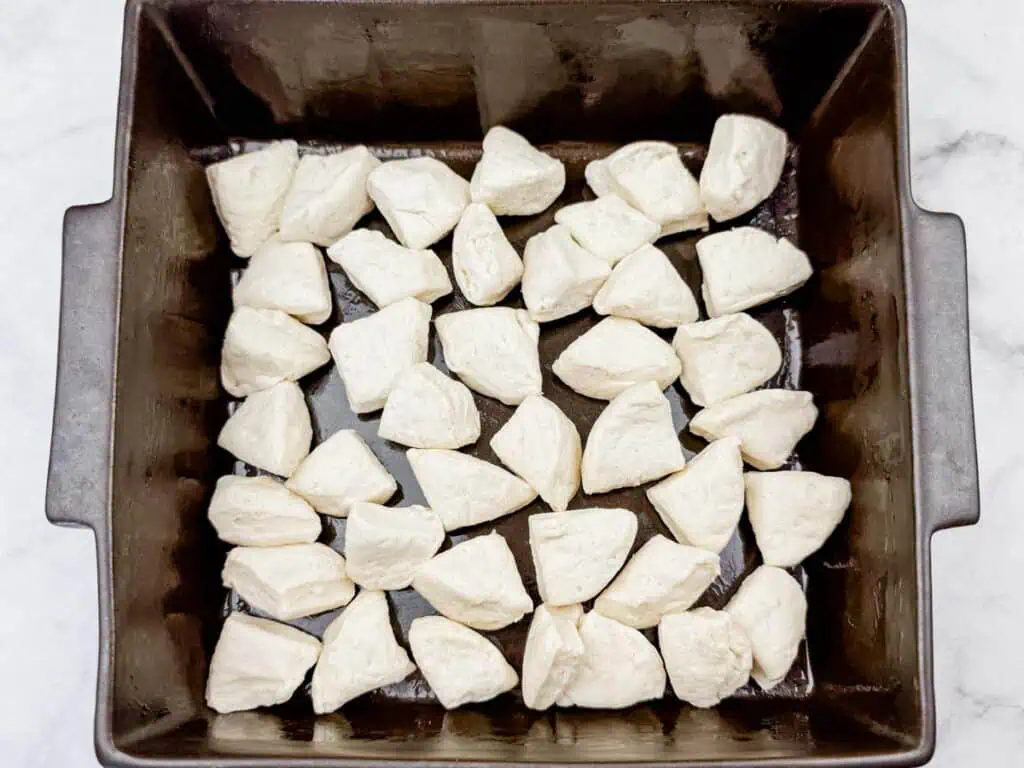 A square baking pan filled with evenly spaced, uncooked biscuit dough pieces, arranged on a white surface.