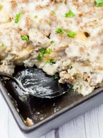 A baking dish with a creamy casserole topped with ground meat and chopped parsley; a spoon rests in one corner, showing a serving removed.