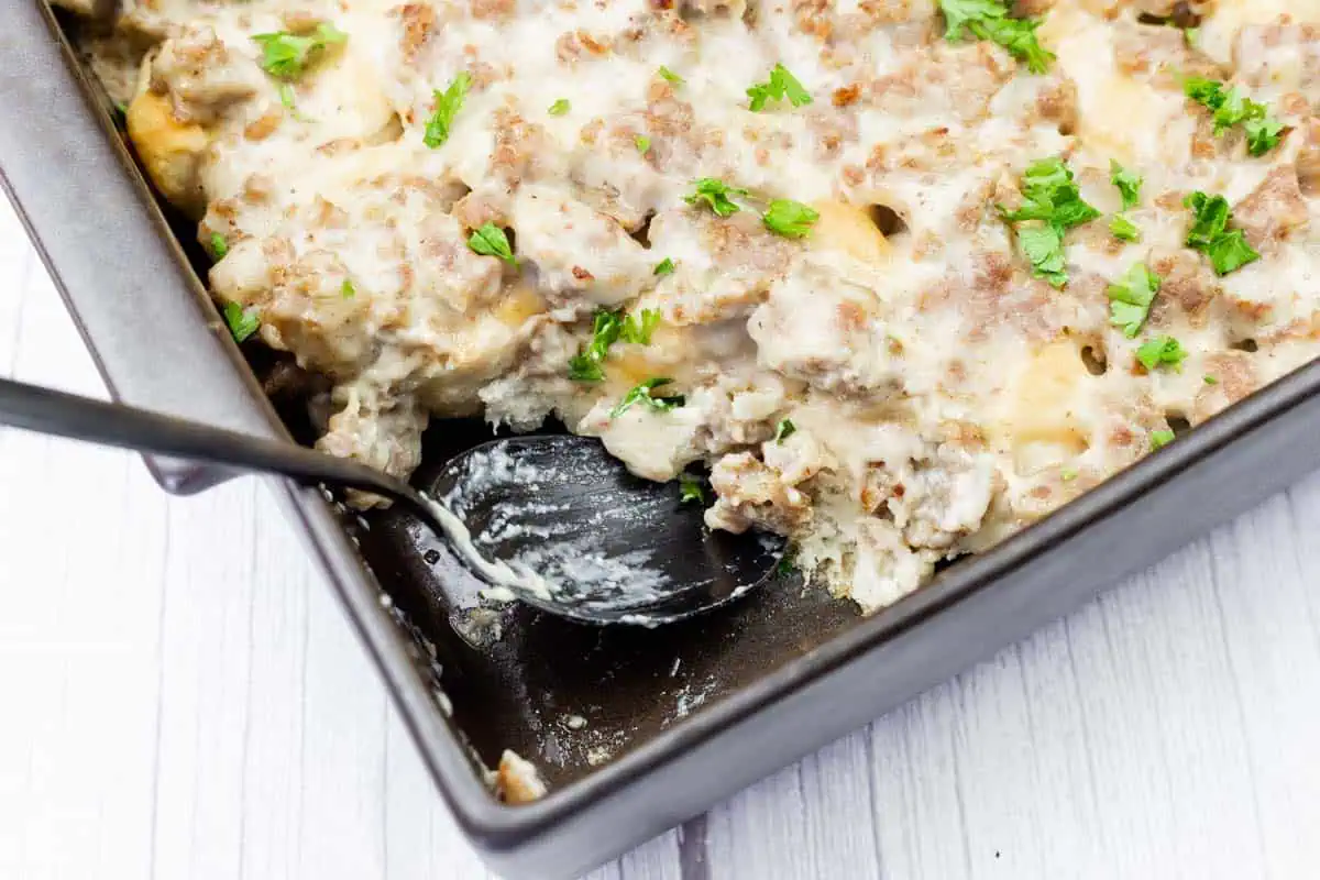 A baking dish of sausage and biscuit casserole topped with creamy gravy and chopped parsley, with a serving spoon partially in the dish.