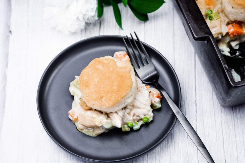 A black plate with a serving of chicken pot pie topped with a biscuit, a fork, and a casserole dish with more pot pie partially visible.