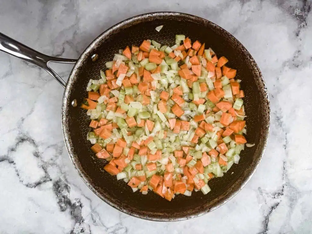 Pan with diced carrots, celery, and onions sautéing on a marble countertop.