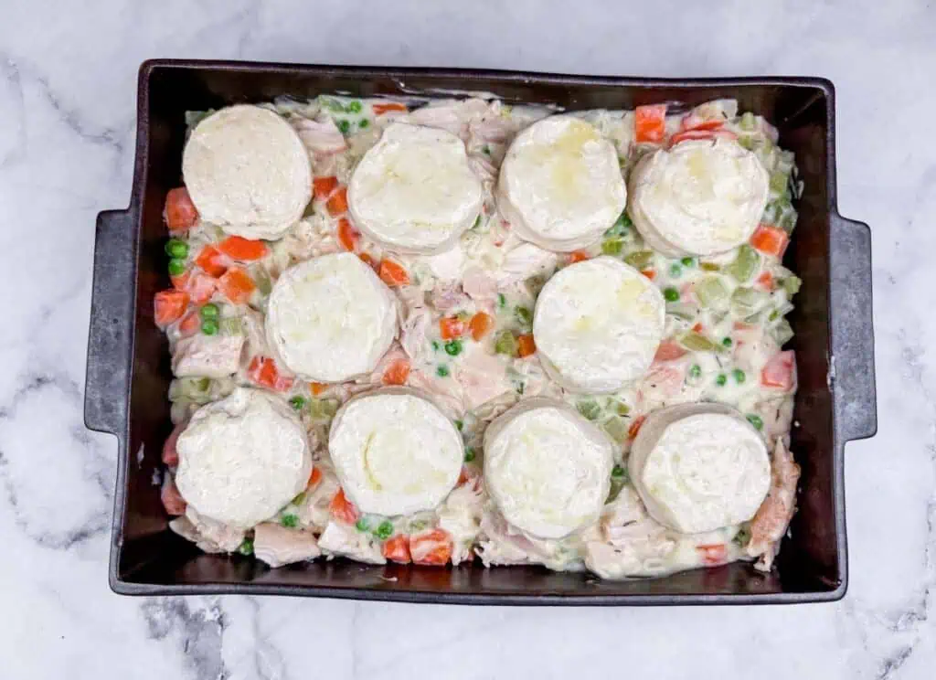 A baking dish filled with a mixture of vegetables and chicken in a creamy sauce, topped with nine uncooked biscuit dough rounds.