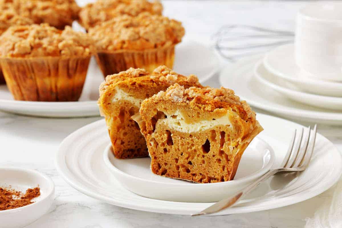 A pumpkin muffin with a cream cheese center is cut in half on a white plate, with more muffins, a fork, and dishes in the background.
