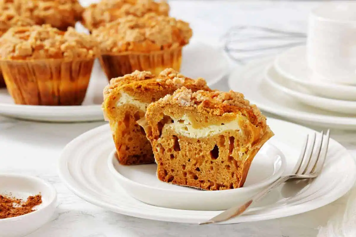 A pumpkin muffin with a cream cheese center is cut in half on a white plate, with more muffins, a fork, and dishes in the background.