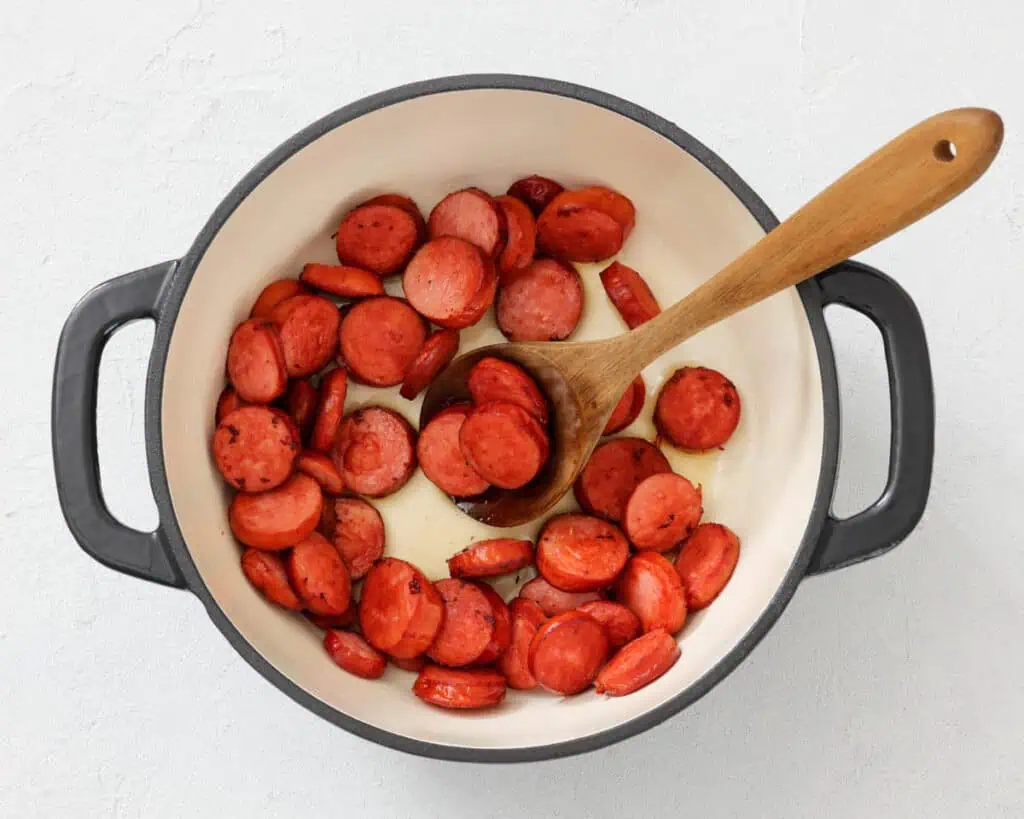 Sliced sausage pieces are being cooked in a white pot with a wooden spoon on a light surface.