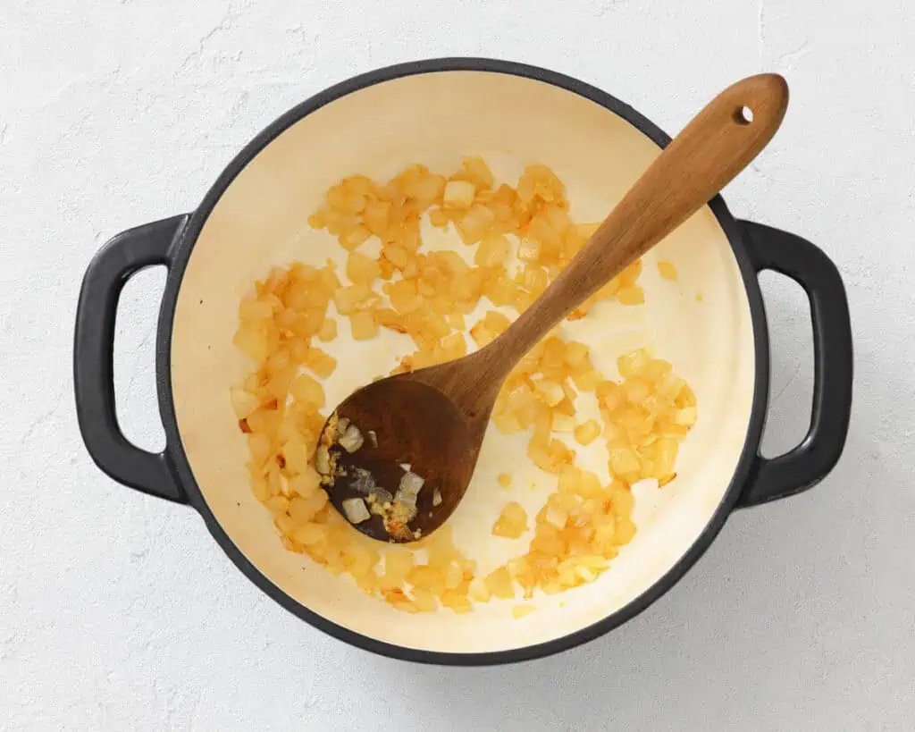 Diced onions sautéing in a white pot with a wooden spoon, placed on a light-colored surface.