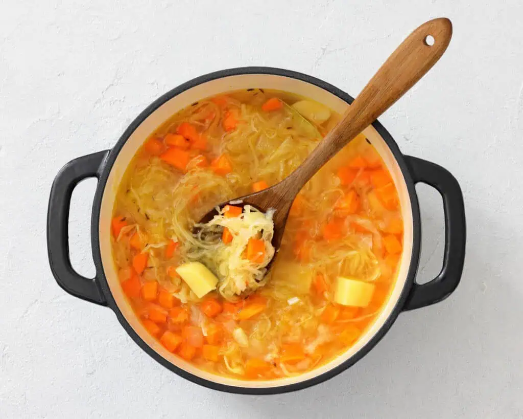 A pot of vegetable soup with carrots, potatoes, and shredded cabbage is being stirred with a wooden spoon.