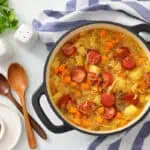 A pot of Chef Jenn’s Sauerkraut Soup with Kielbasa sits on a striped towel next to wooden spoons, bowls, fresh herbs, and salt and pepper shakers.