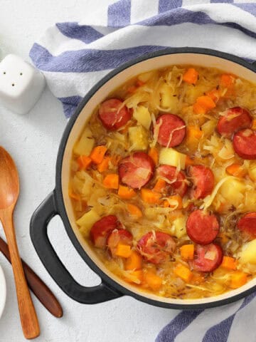 A pot of Chef Jenn’s Sauerkraut Soup with Kielbasa sits on a striped towel next to wooden spoons, bowls, fresh herbs, and salt and pepper shakers.