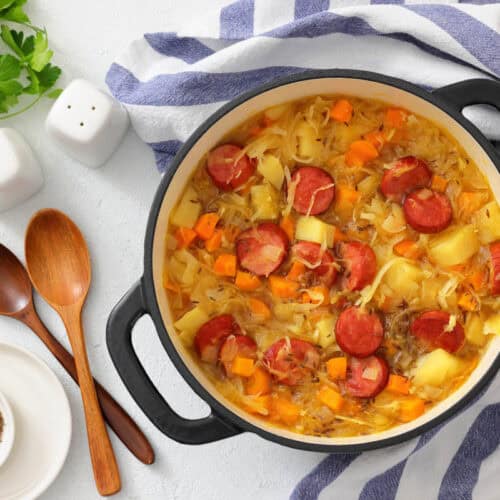 A pot of Chef Jenn&rsquo;s Sauerkraut Soup with Kielbasa sits on a striped towel next to wooden spoons, bowls, fresh herbs, and salt and pepper shakers.