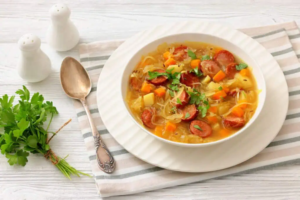 A bowl of vegetable and sausage soup garnished with parsley, placed on a striped cloth next to a spoon, a fork, salt and pepper shakers, and a bunch of fresh parsley.