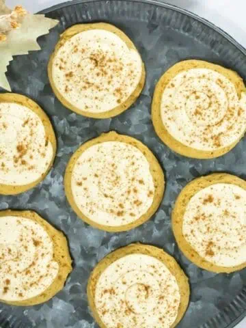 Crumbl Caramel-Pumpkin Cookies are arranged on a metal tray, accompanied by fall-themed decorations and cinnamon sticks.with brown spice. Decorative fall elements and a drink are nearby.