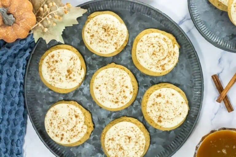 Crumbl Caramel-Pumpkin Cookies are arranged on a metal tray, accompanied by fall-themed decorations and cinnamon sticks.with brown spice. Decorative fall elements and a drink are nearby.