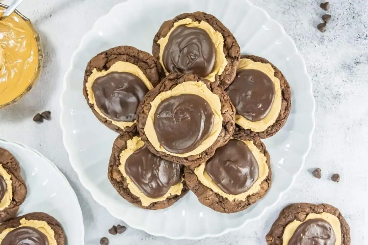 A white plate holds several chocolate cookies topped with a layer of peanut butter and a swirl of chocolate ganache.
