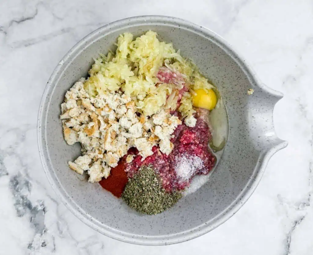 A mixing bowl containing soaked bread, ground beef, ground pork, onion, egg, parsley, mustard, marjoram, salt, pepper, and paprika on a marble surface.