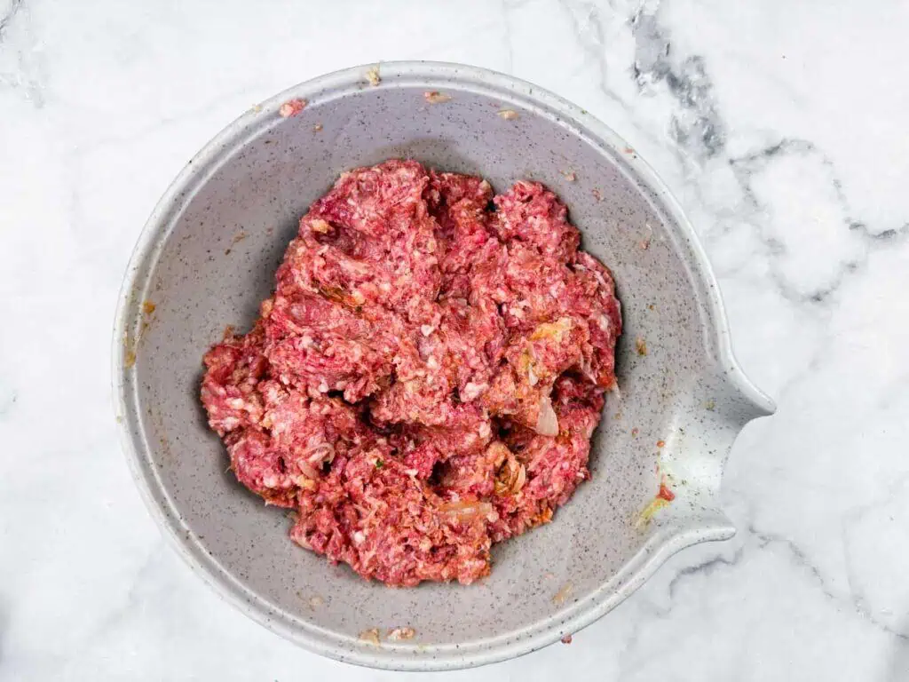 A gray mixing bowl filled with raw ground meat mixture sits on a marble countertop.