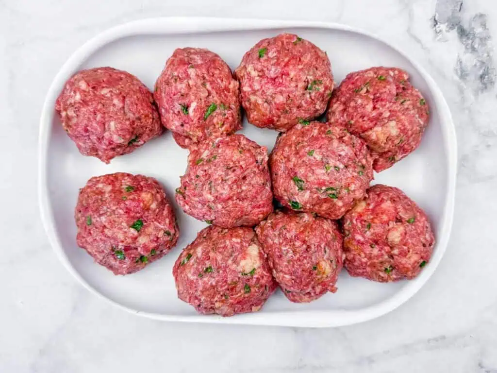 Ten raw meatballs made from ground meat and herbs are arranged on a white oval plate set on a marble surface.