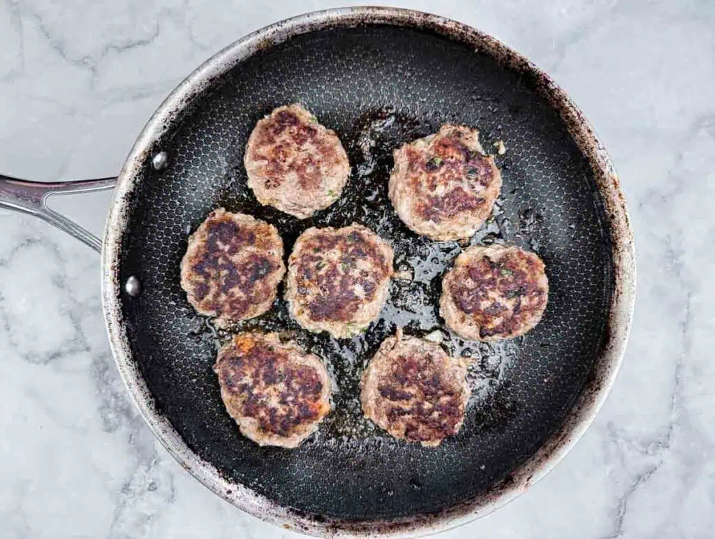 Seven browned patties cooking in a black frying pan on a light gray countertop.