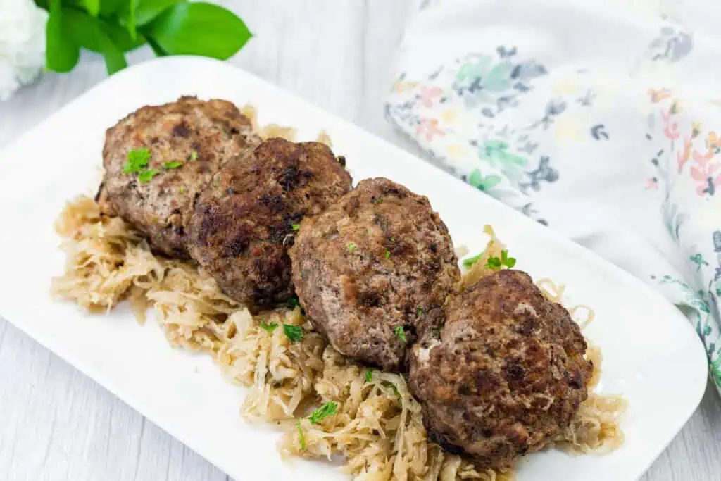 Four browned meat patties are served on a bed of shredded cabbage or sauerkraut on a white rectangular plate. Floral fabric and greenery are in the background.