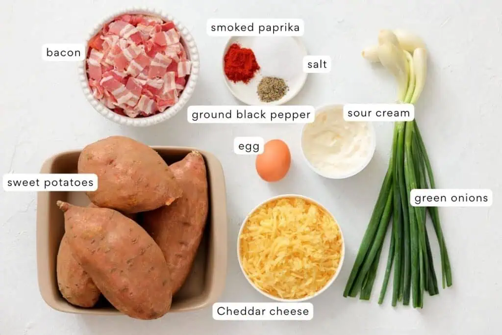 Ingredients for a recipe are arranged on a white surface, including sweet potatoes, bacon, cheddar cheese, green onions, sour cream, egg, smoked paprika, salt, and pepper. Labels identify each item.