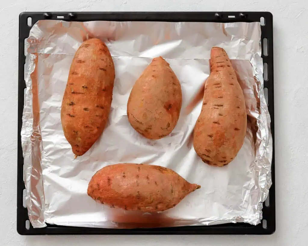 Four whole sweet potatoes placed on a foil-lined baking tray, ready for baking.