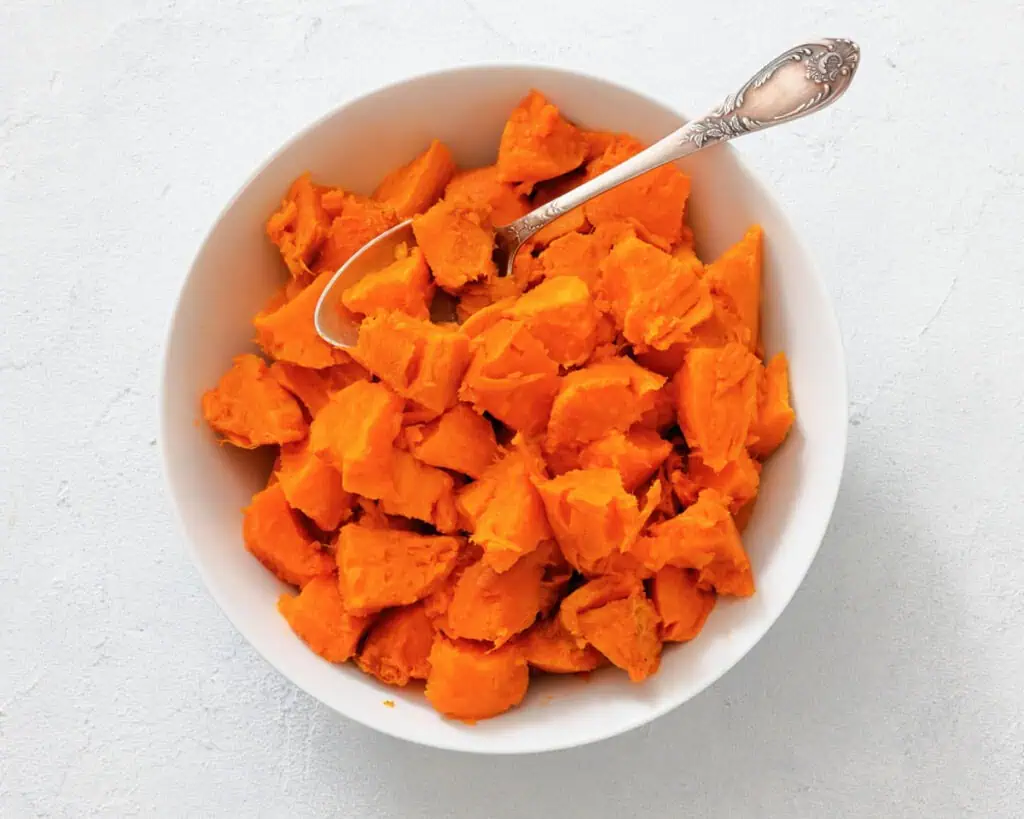 A white bowl filled with chunks of cooked sweet potato, with a silver spoon resting inside.