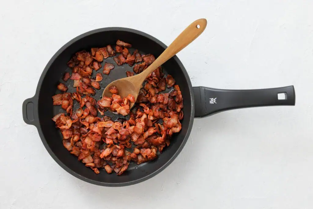 Chopped bacon pieces cooking in a black frying pan with a wooden spoon on a light surface.