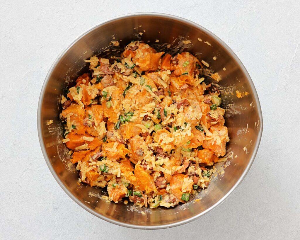 A stainless steel bowl containing a mixed salad with sweet potatoes, shredded cheese, chopped herbs, bacon, and green onions.