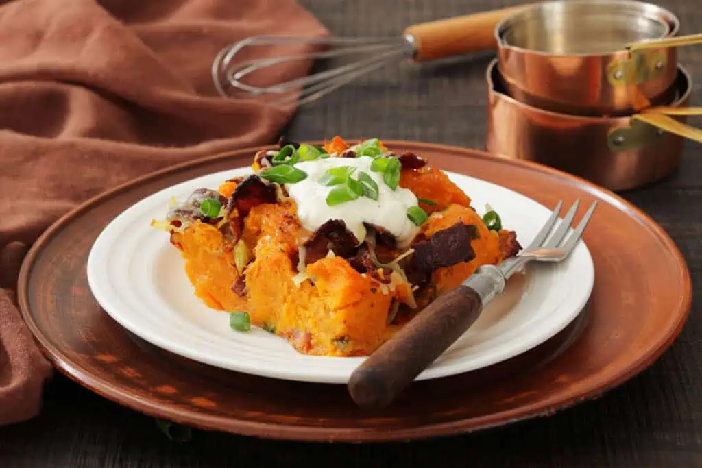 A plate of loaded sweet potato casserole topped with sour cream, bacon, cheese, and chopped green onions sits on a wooden charger with a fork, whisk, and measuring cups nearby.
