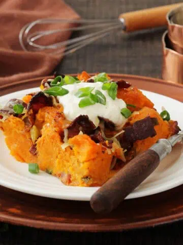 A plate of loaded sweet potato casserole topped with sour cream, bacon, cheese, and chopped green onions sits on a wooden charger with a fork, whisk, and measuring cups nearby.