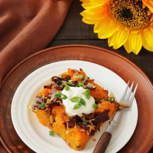 A plate with a serving of loaded sweet potato casserole topped with sour cream, chopped green onions, and bacon, with a fork beside it; sunflowers are in the background.