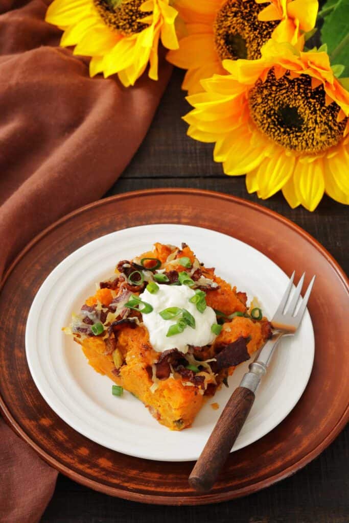 A plate with a serving of loaded sweet potato casserole topped with sour cream, chopped green onions, and bacon, with a fork beside it; sunflowers are in the background.