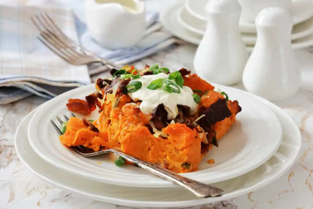 A serving of baked sweet potato casserole topped with sour cream, chopped green onions, bacon, and cheese on a white plate with a fork.