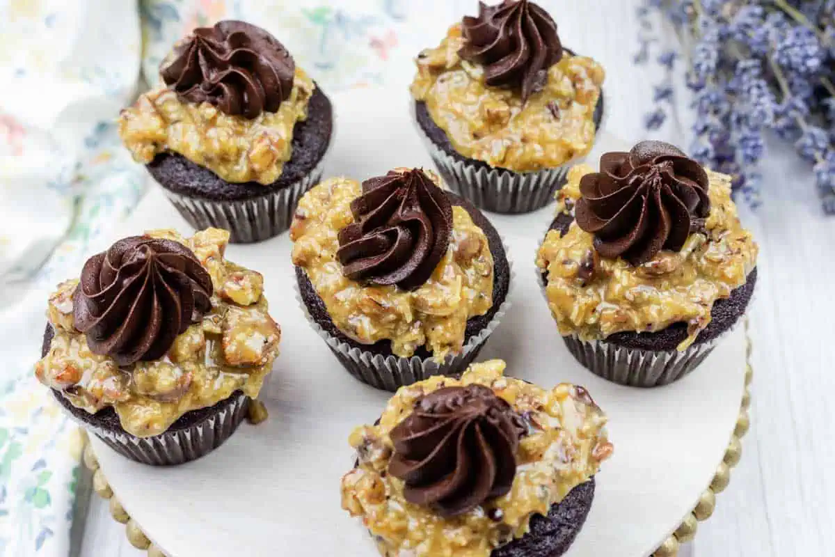 Six German chocolate cupcakes topped with a layer of caramel pecan coconut frosting and a swirl of chocolate frosting, displayed on a round wooden board.
