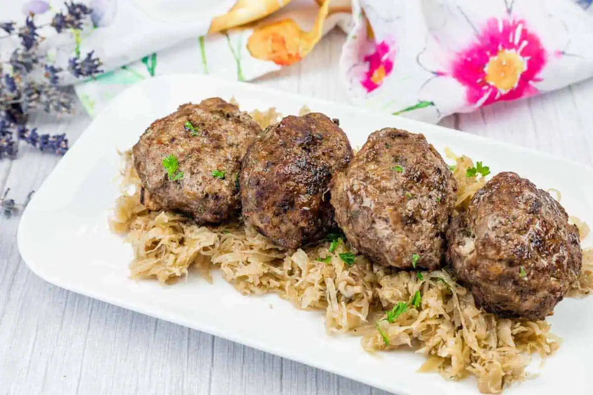 Four German meat patties are served on a bed of sauerkraut, garnished with parsley, on a rectangular white plate with a floral napkin in the background.