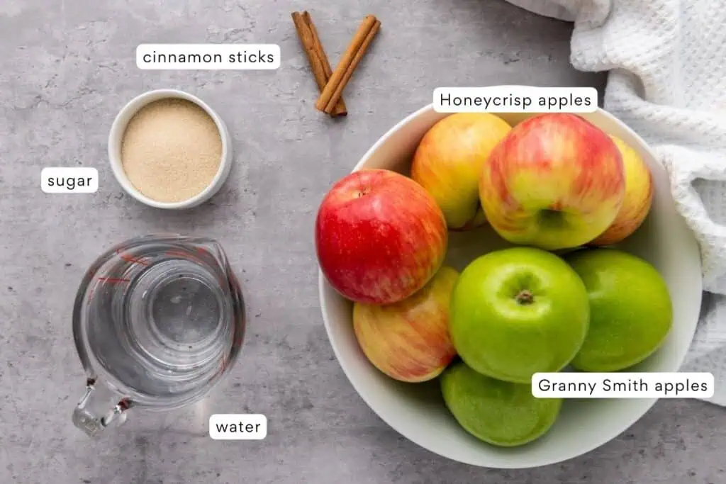 A bowl with Honeycrisp and Granny Smith apples, a small bowl of sugar, a measuring cup of water, and two cinnamon sticks on a gray surface.