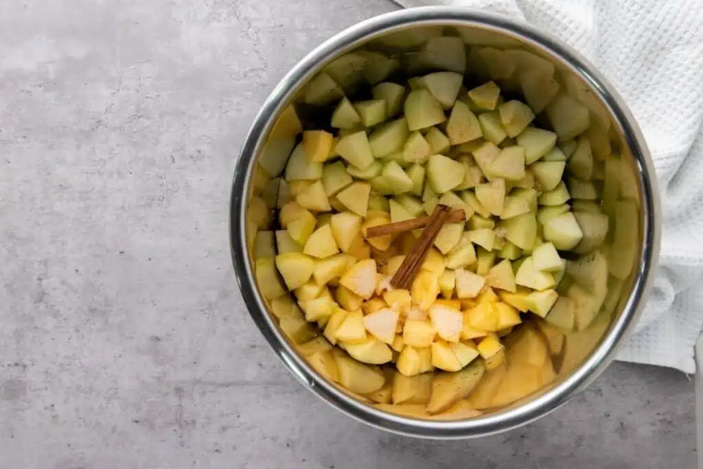 Chopped apples and pears with a cinnamon stick and sugar in a metal pot, placed on a gray countertop next to a white towel.