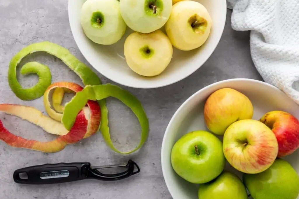 Two bowls of apples, one with peeled apples and one with unpeeled apples, sit on a gray surface beside apple peels, a peeler, and a white textured cloth.