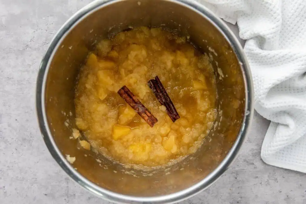 A stainless steel pot containing chunky applesauce with two cinnamon sticks, placed on a light gray surface next to a white textured cloth.