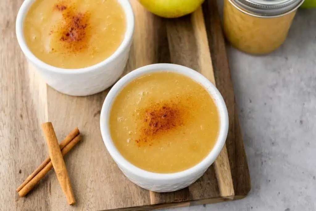 Two small white bowls of applesauce topped with cinnamon sit on a wooden board next to cinnamon sticks, an apple, and a jar of applesauce.