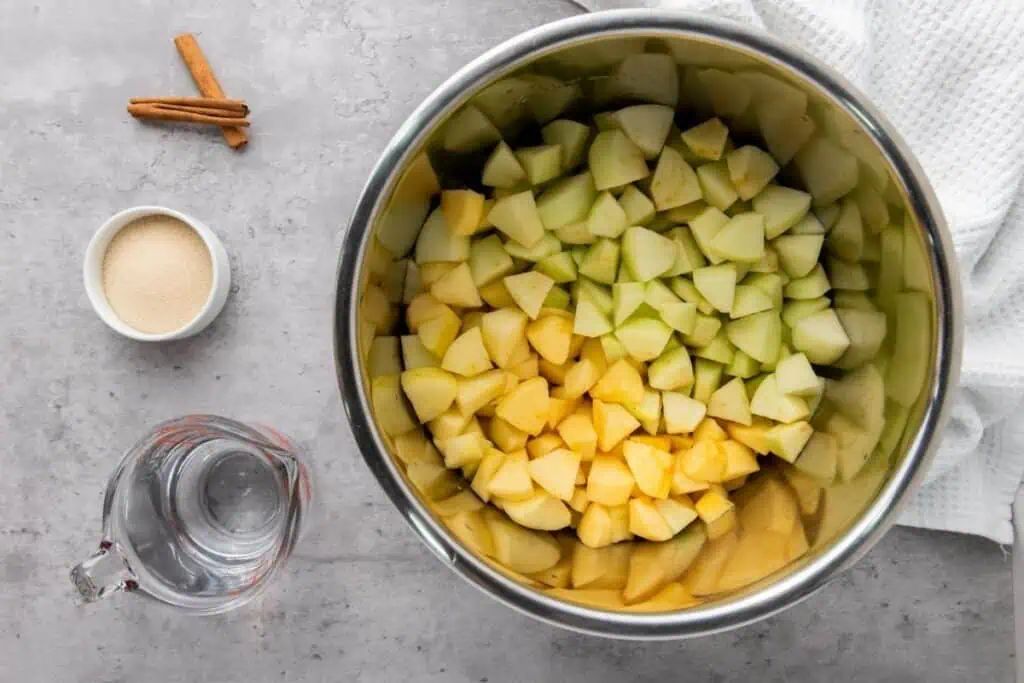 A metal bowl filled with chopped apples sits on a gray countertop next to a small bowl of sugar, a glass measuring cup of water, and two cinnamon sticks.