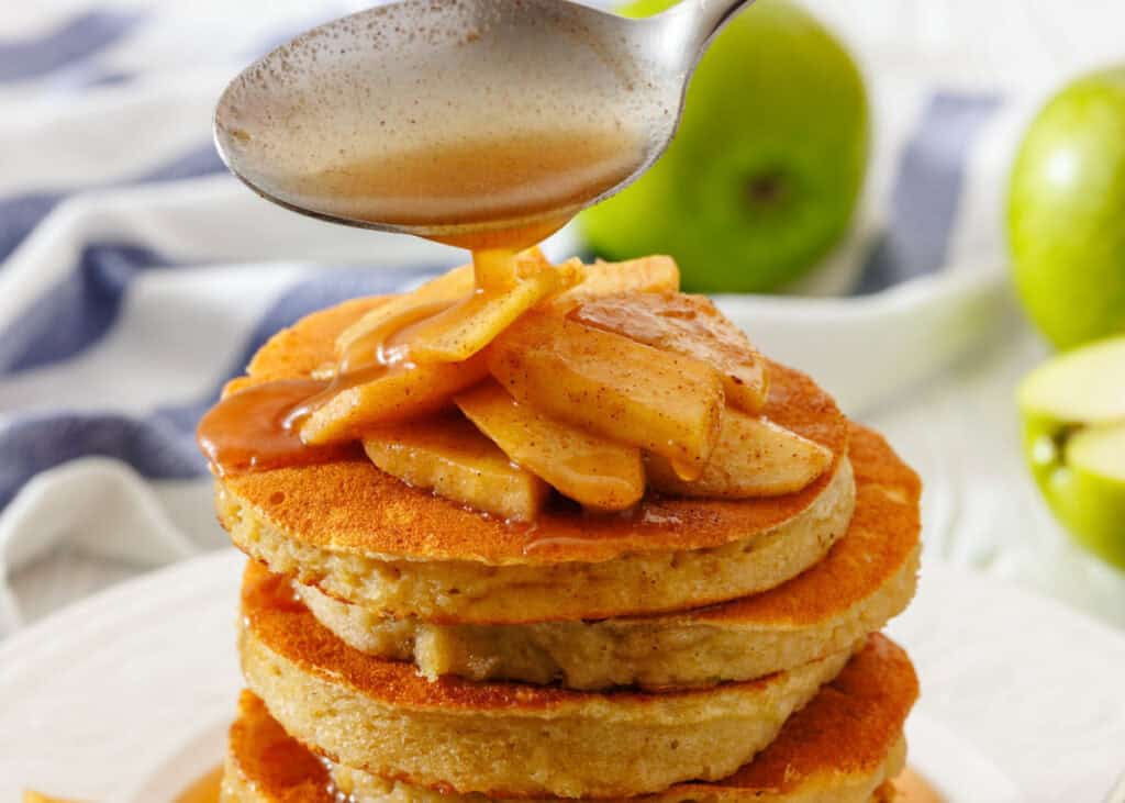 A stack of pancakes topped with cooked apple slices is being drizzled with syrup from a spoon, with green apples in the background.