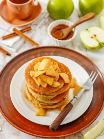 A stack of pancakes topped with cooked apple slices sits on a white plate with a fork. Green apples, cinnamon sticks, and a cup are in the background.