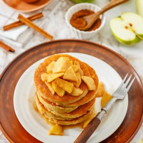 A stack of pancakes topped with cooked apple slices sits on a white plate with a fork. Green apples, cinnamon sticks, and a cup are in the background.