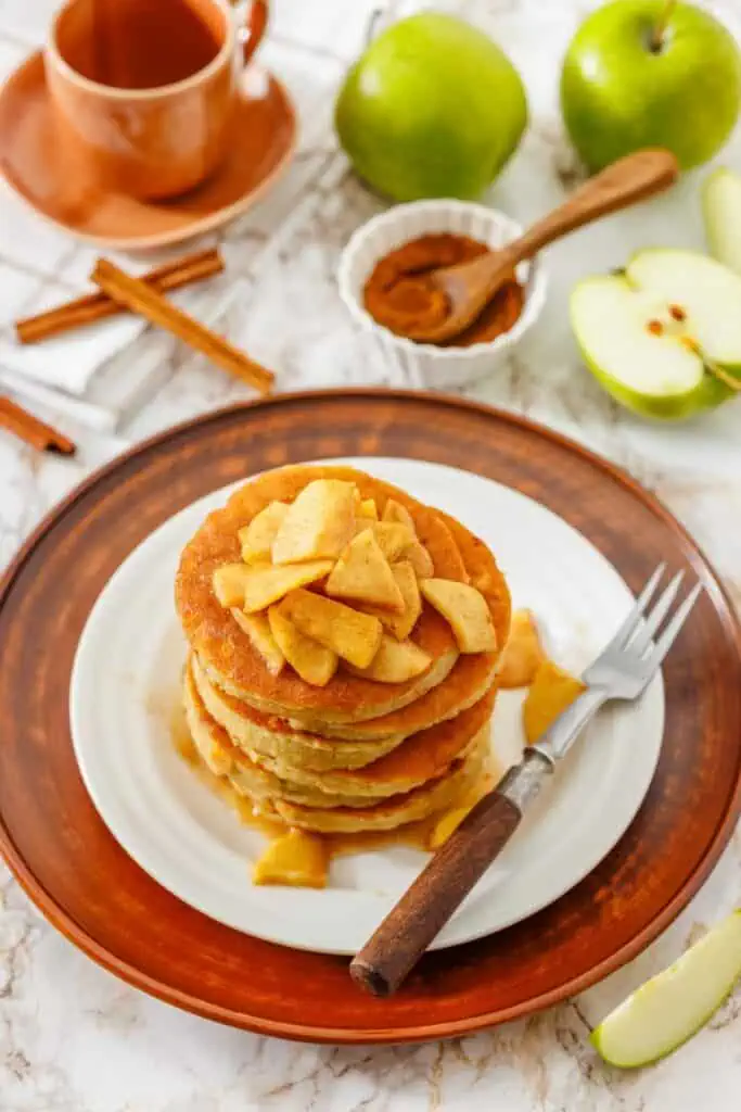 A stack of pancakes topped with cooked apple slices sits on a white plate with a fork. Green apples, cinnamon sticks, and a cup are in the background.
